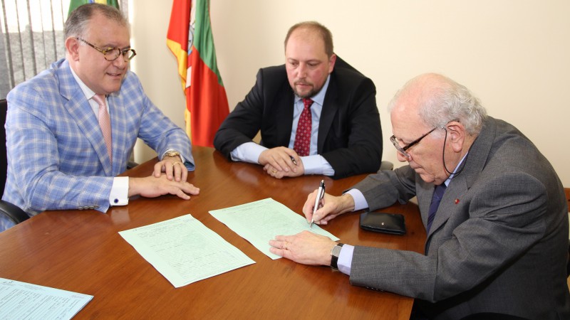Ivo Nesralla, assinando o documento, secretário Alessandro Barcellos e Alberto Beltrame