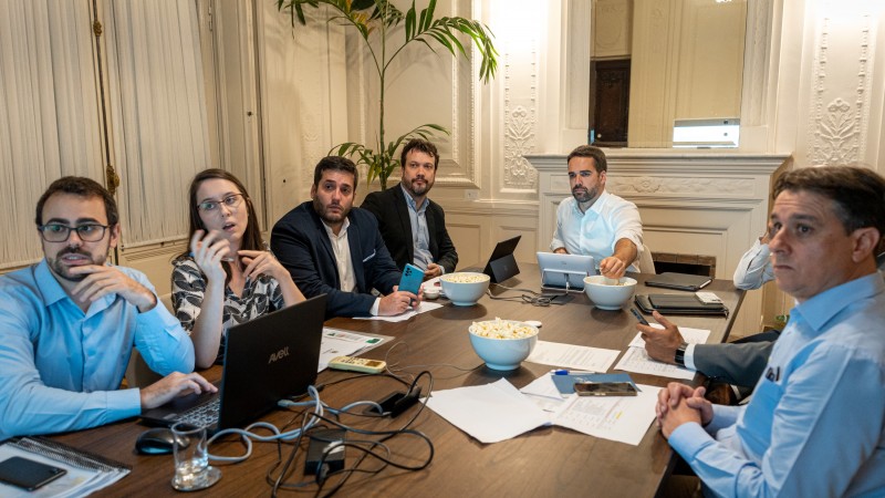 Governador Eduardo Leite, de camisa branca, sentado diante da cabeceira de mesa de reuniões. Ao redor, estão o chefe da Casa Civil, Artur Lemos, e integrantes da assessoria técnica do gabinete. Sobre a mesa, notebooks, tablets, papéis e tigelas com pipoca.