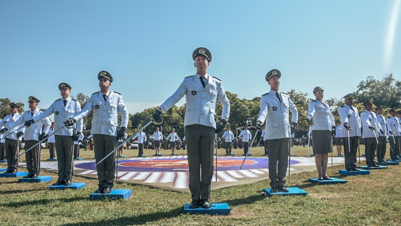 PMs com farda composta por gandola branca e calça ou saia ocre, todos com cap de cobertura, aparecem alinhados em fila empunhando as espadas de oficias. Cada um deles está sobre um suporte quadrado de cor azul, no gramado do estádio do complexo de Ensino da BM.