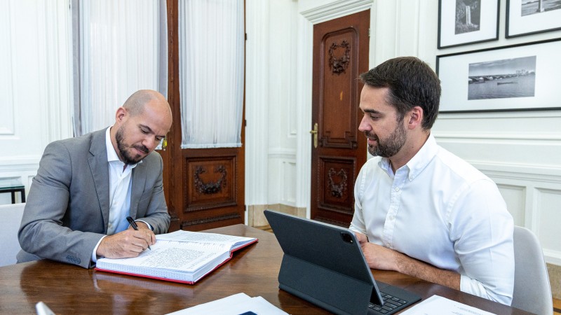 Economista Pedro Capeluppi, de terno cinza e camisa branca, assina livro de posse como secretário de Parcerias e Concessões. Ele está sentado diante de uma mesa ao lado do governador Eduardo Leite, também sentado, de camisa branca, com um tablet a sua frente.