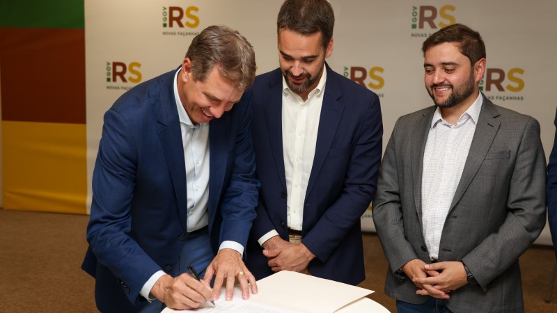 Ernani Polo assina termo de posse como secretário de Desenvolvimento Econômico, ao lado do governador Eduardo Leite e do vice-governador Gabriel Souza. Os três estão em pé diante de uma mesa. Todos usam camisa branca. Leite e Polo vestem blazers azuis e Gabriel, cinza.