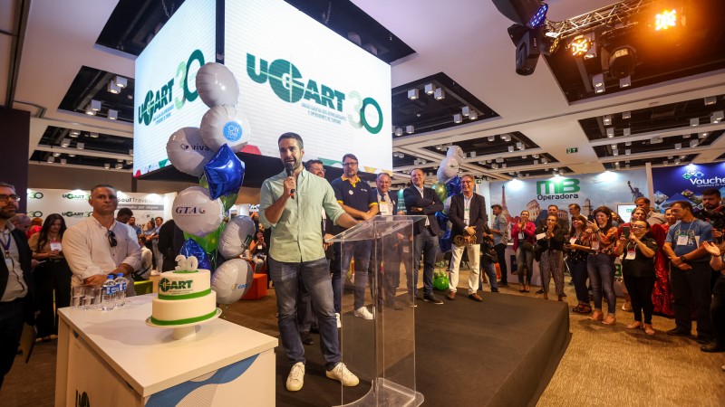 Eduardo Leite, em pé, de camisa verde claro, calça jeans e tênis branco, fala segurando um microfone diante de um púlpito acrílico, sobre um palco com outras pessoas, acima do qual há telões suspensos formando um quadrado. Ao lado do palco, uma bancada com um bolo de aniversário de 30 anos da Ugart e balões de gás hélio. Ao redor, público presente.