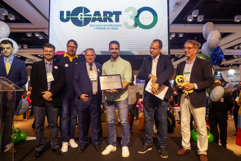 Eduardo Leite, em pé, de camisa verde claro, calça jeans e tênis branco, segura uma placa de homenagem. Ao lado dele, à esquerda e à direita, outros homens de terno. Um deles segura uma grande chave dourada. Ao fundo, um telão com a logo Ugart 30 anos.