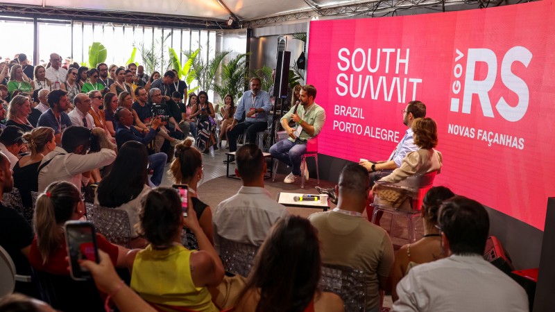 Em um palco diante de um telão com fundo rosa e as logos do South Summit Brazil e do governo do RS, aparecem sentados em cadeiras o governador Eduardo Leite, o secretário da SJCDH Mateus Wesp, e a secretária da Educação, Raquel Teixeira. Diante do palco, grande plateia.