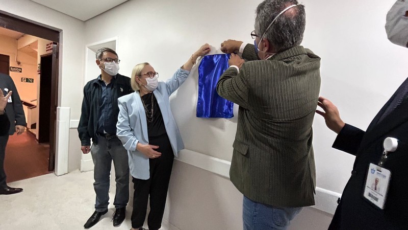 Secretária Arita Bergmann, em pé, de máscara, blazer azul claro, blusa e calça preta, descerra cortina de cetim azul que cobre a placa de inauguração da nova ala de saúde mental em hospital de Rio Grande. Também puxa a cortina um homem de blazer bege e calça jeans.