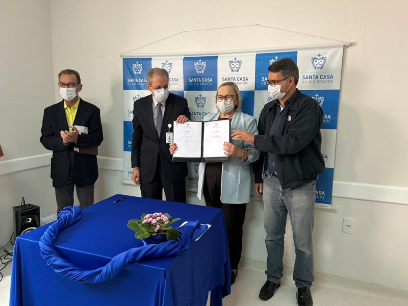 Secretária Arita Bergamann mostra termo assinado para adesão da Santa Casa do Rio Grande à rede estadual de urgência e emergência, que assegura repasse mensal de R$ 500 mil. Ao lado dela, o prefeito de Rio Grande, Fábio Branco, e o diretor do hospital. Atrás deles, um banner quadriculado em azul e branco com logos da Santa Casa.