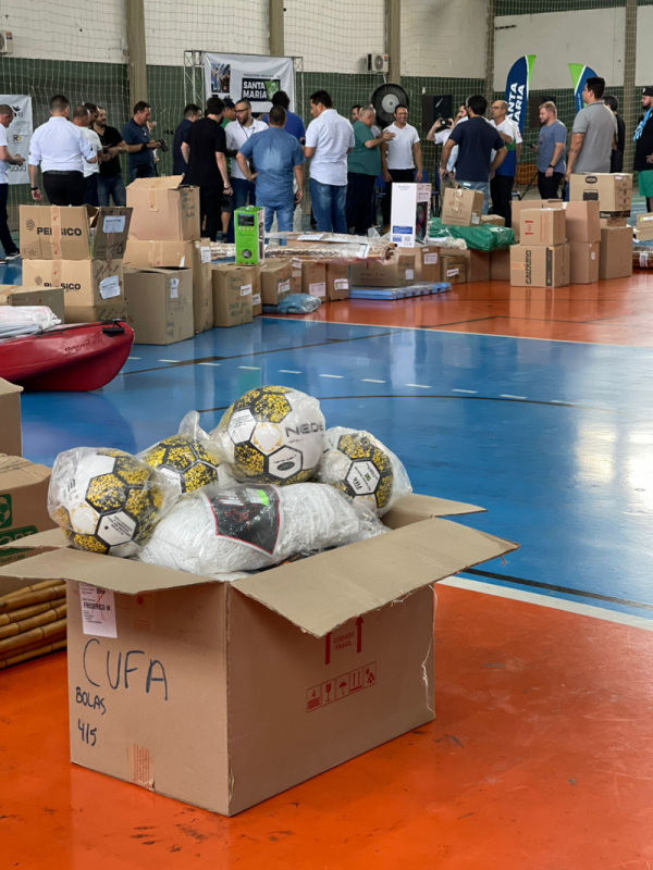 Caixa de papelão com a inscrição CUFA, contendo bolas de futebol, aparece em primeiro plano no piso de um ginásio de esportes. Ao fundo, várias outras caixa e pessoas reunidas.