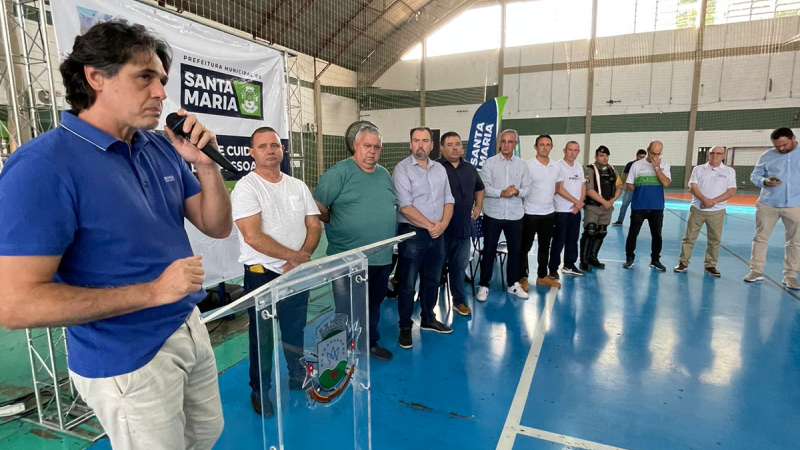 Secretário Danrlei, de camisa polo azul e calça jeans branca, em pé, fala segurando um microfone diante de um púlpito de acrílico transparente. Ao lado dele, vários homens lado a lado. Eles estão em um ginásio de esportes. Ao fundo, banner com a logo da prefeitura de Santa Maria.