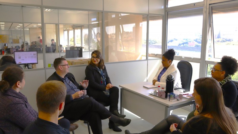 Em uma sala com divisórias transparentes na lateral e janelas ao fundo, a secretária adjunta da SJCDH, Caroline Moreira, está sentada diante de uma mesa. É uma mulher negra, vestindo blusa azul e casaco branco. Diante dela, dois homens e quatro mulheres participam da reunião. Um dos homens, de casaco preto e camisa azul, dialoga com a secretária.