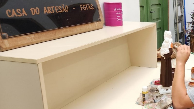 Sobre um balcão bege claro, um quadro negro com bordas de madeira, no qual está escrito em marrom "Casa do Artesão - FGTAS". Ao lado dele, uma lata rosa. Em nível abaixo do balcão, uma escultura em gesso de um santo é pintada por um pincel empunhado pela mão de um dos internos da Fase que conquistou a carteira do Artesão. Ele não aparece na cena para não ser identificado, em respeito ao ECA.