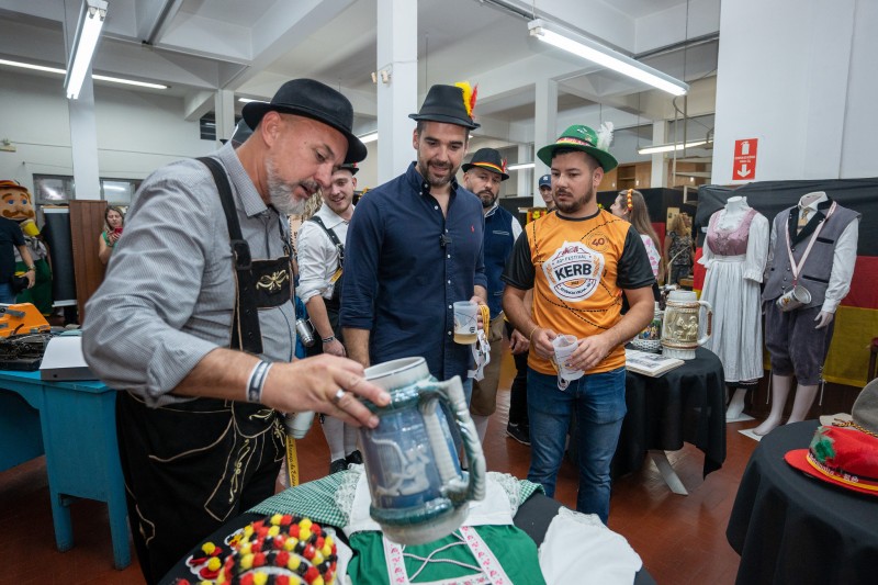 Um homem vestindo camisa cinza e suspensório preto e um chapéu preto, segura um grande caneco de chopp sobre uma mesa. Ao lado, em pé, o governador Eduardo Leite, de camisa azul escura e segurando um caneco pequeno transparente de chopp, e vestindo um chapé preto com uma pena amarela e vermelha, observa, acompanhado de outra pessoa vestindo a camiseta do 40º Kerb de Estância Velha. Ao fundo, manequins com trajes típicos da imigração alemã, no interior do Museu do Kerb.