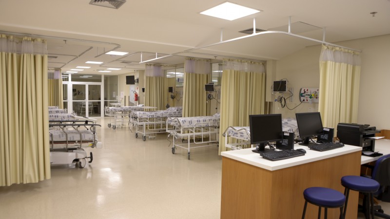 Imagem mostra uma grande sala de hospital com camas separadas por cortinas amarelas, em primeiro plano, um balcão com dois computadores e uma impressora. Atrás dele, duas banquetas azuis. É o espaço da nova emergência do Hospital de Clínicas de Passo Fundo, que passou de 900 m2 para 2.100 m2.
