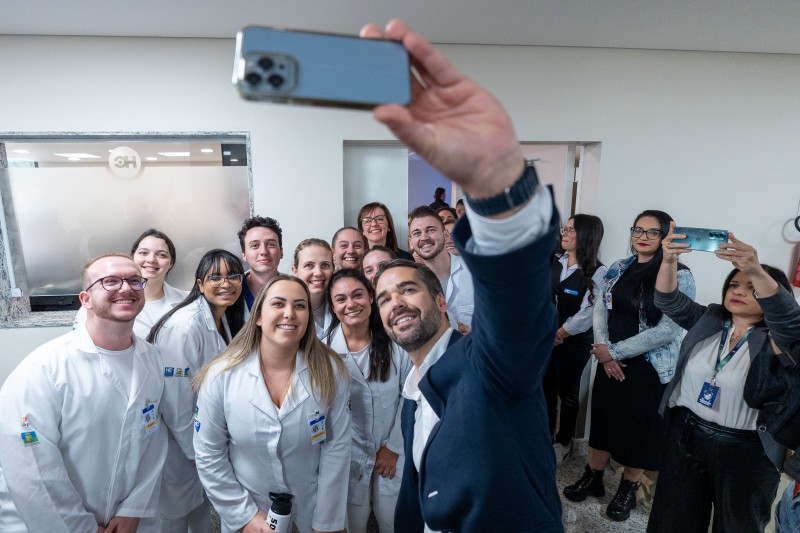 O governador Eduardo Leite, de casaco azul e camisa branca, segura com a mão esquerda um celular no alto para tirar uma selfie com um grupo de funcionários do Hospital de Clínicas de Passo Fundo, todos vestindo jalecos brancos.
