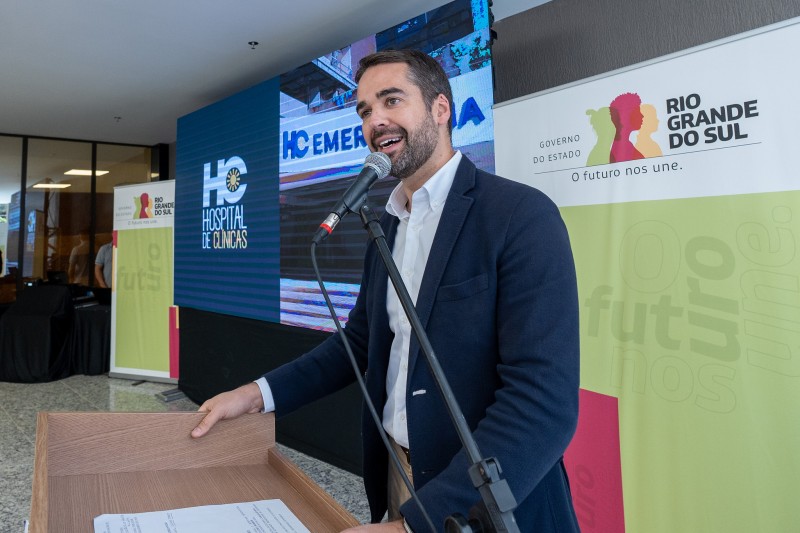 Eduardo Leite, de casaco azul e camisa branca, fala diante de um microfone em um pedestal, em frente a um púlpito de madeira. Ao fundo, telão com a marca do Hospital de Clínicas e banners com a marca do governo do Estado.