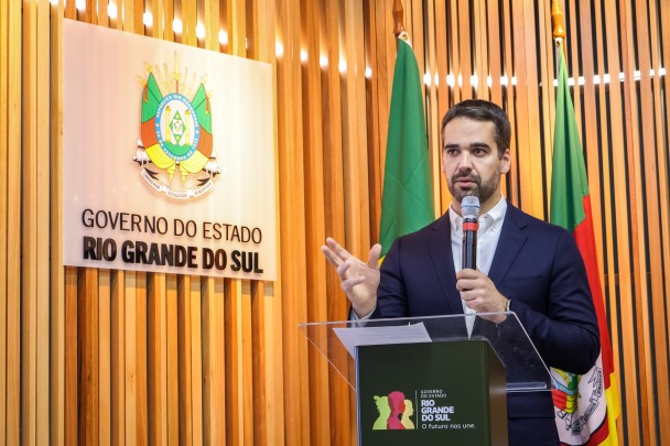 O governador Eduardo Leite fala segurando um microfone diante de um púlpito. Atrás dele, bandeiras do Brasil e do RS. Ao lado, brasão do RS em uma peça de acrílico afixada em uma parede de ripas de madeiras.