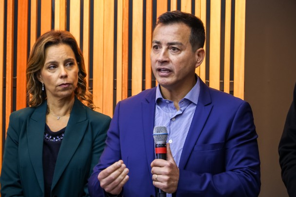 Secretário da Segurança Pública, Sandro Caron, fala ao microfone. Ele está de casaco azul marinho e camisa lilás. Ao lado dele, uma mulher de casaco verde, blusa preta, cabelos loiro escuto, observa.