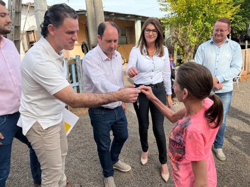 O secretário Fabrício Perucchin, homem branco, de cabelo preto ondulado, camisa branca e calça marrom, entrega uma chave nas mãos de uma moradora de Venâncio Aires, Adriana Silveira, uma mulher de cabelo castanho e camiseta rosa. Outros três homens e uma mulher, todos de camisas claras e calça jeans, acompanham o momento, que acontece na rua, em frente a casa da moradora, no bairro Battisti.