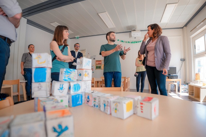 La foto, en un salón de clases, muestra en primer plano una mesa con un montón de blocs de papel con dibujos de niños.  De fondo, el Vicegobernador Eduardo Leite habla sobre el proyecto de escuela pública de tiempo completo con dos mujeres.  Otras personas en la habitación están mirando.