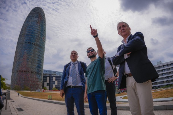 En una plaza, el vicegobernador Gabriel Souza apunta hacia arriba con su brazo izquierdo hacia el horizonte.  A su lado, dos hombres con traje miran en la dirección que señala.  Al fondo, a la izquierda, un gran edificio con fachada de espejos se destaca contra el cielo nublado.