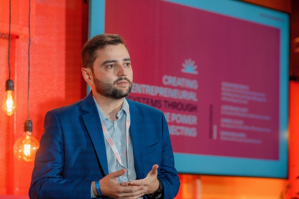 El Teniente Gobernador Gabriel Souza, con traje azul marino y camisa celeste, con un lazo distintivo al cuello.  Tiene el pelo castaño corto y una barba recortada.  En el fondo, a la izquierda de Gabriel, dos bombillas con cables amarillos cuelgan de cables con el dorso negro.  Más atrás, a la derecha, en una imagen borrosa, una pantalla con una diapositiva de color lila con textos en blanco.