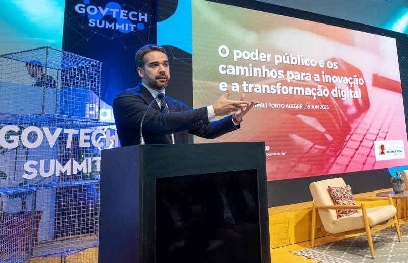 Leite em close, atrás de um púlpito preto à esquerda da foto, gesticulando de tal maneira que a posição das mãos parecem oferecer ao público o conteúdo que aparece escrito em um painel vermelho localizado atrás de si, à direita da foto.