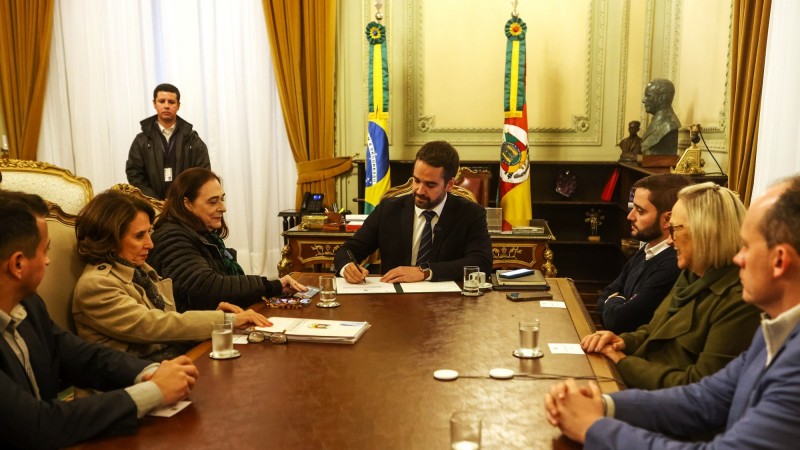 Govenador Eduardo Leite sentado na cabeceira de uma mesa de reunião assinando um termo de parceria com a Unesco para aprimorar a educação gaúcha. Do lado esquerdo há três pessoas, entre elas a representante da Unesco e a secretária da Educação. Do lado direito também aparecem três pessoas à mesa e entre elas está o vice-governador Gabriel Souza.