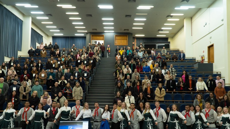 Foto tirada a partir do palco do público no auditório. Todos estão de pé. À frente, estão enfileirados jovens com trajes gaúchos típicos que se apresentaram na ocasião.