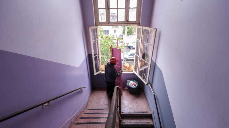 Trabalhador carrega uma porta cor-de-rosa na escadaria da escola, em frente a uma janela