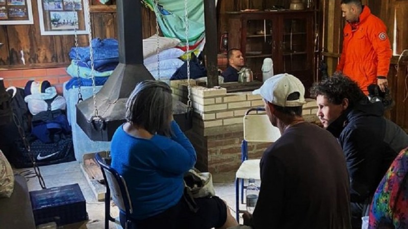 Imagem interna do CTG. Três pessoas conversam de costas e ao fundo dois homens conversam. Do lado superior esquerdo da foto é possível ver materiais que parecem ser colchões e cobertores doados.