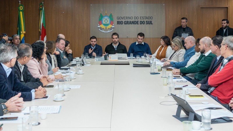 Ao centro Eduardo Leite na cabeceira de uma grande mesa branca. Na cabeceira duas pessoas estão ao lado do governador. Uma delas é o vice-governador que está à esquerda. Ao longo da mesa, dos dois lados, estão os secretários do governo.