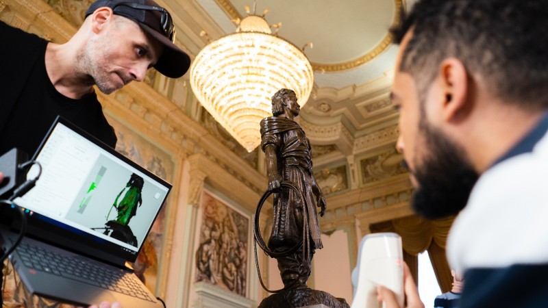 Um pesquisador segura, à esquerda, um notebook em que aparece a figura digitalizada, no caso, a estátua do Laçador; à direita, outro pesquisador realiza o escaneamento