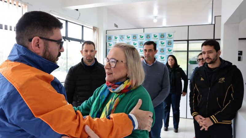 Em primeiro plano à esquerda o prefeito de Caraá olha a secretária da Saúde de frente. Ele segura nos ombros dela e ela segura os braços dele. Ao fundo, pessoas assistem à cena.