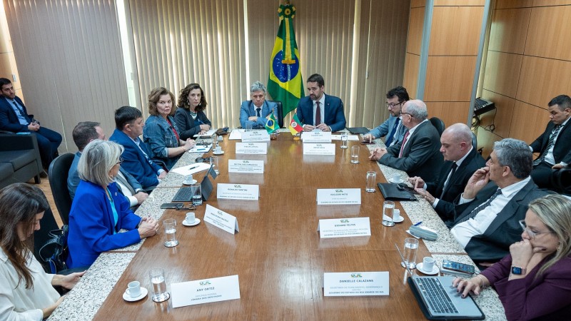 Em uma mesa comprida preta Leite está dividindo a cabeceira com o ministro. Eles estão mais distantes na imagem. Podem ser vistas várias pessoas que participam da reunião dos dois lados da mesa.