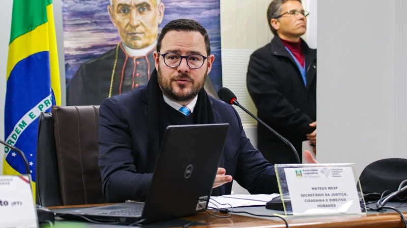 Secretário Mateus Wesp em primeiro plano, sentado atrás de uma bancada e diante de um microfone, apresentando o PL.