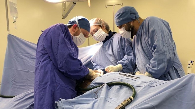 Imagem do centro cirúrgico. Há três médicos realizando o procedimento. Não se vê a paciente, pois ela está toda coberta por panos azuis, utilizados para criar o campo cirúrgico.