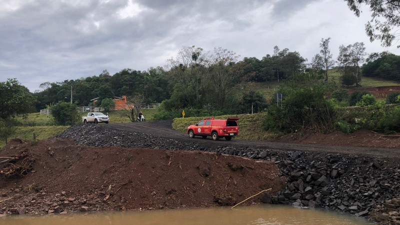 Um acesso asfáltico, rodeado por terra e lama, é atravessado por um veículo vermelho, num sentido, enquanto uma caminhonete prateada, no sentido contrário aguarda sua vez