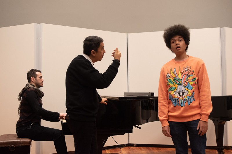 Um homem toca piano ao fundo, enquanto um professor, com a mão elevada, orienta o canto de um jovem