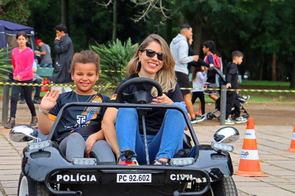 Uma policial civil, branca, loira, de óculos escuro, camiseta preta e calça jeans, pilota um mini buggy da Escolinha de Trânsito da Polícia Civil. Na carona, ela leva uma menina negra, cabelo castanho, camiseta preta com estampas, e calça legging cinza, que acena para o fotógrafo. Ambas sorriem. Ao fundo, é possível ver cones de sinalização de trânsito e outras pessoas caminhando no parque da redenção.