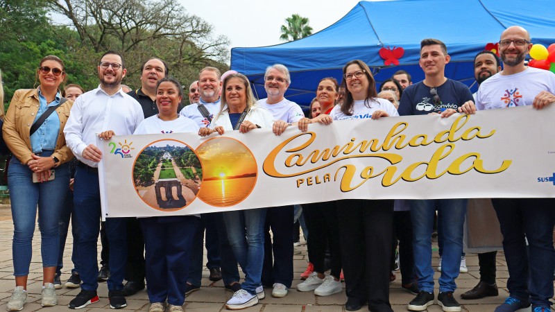 Autoridades e representantes de instituições participantes da Semana Estadual de Prevenção ao Uso de Drogas aparecem lado a lado segurando uma faixa branca com duas fotos em formato de círculo, uma com imagem aérea do Parque de Redenção e outra com imagem do pôr-do-sol no Guaíba. Ao lado das imagens, o texto "Caminhada pela Vida" escrito em letras cursivas na cor amarelo.