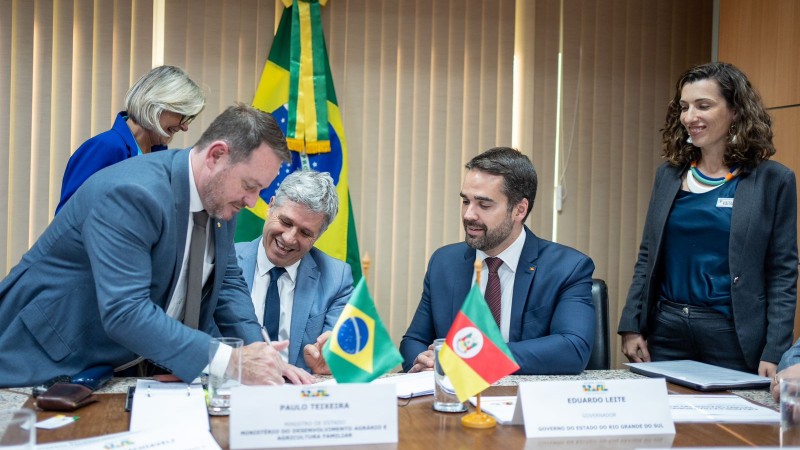 Numa sala fechada, com a bandeira do Brasil ao fundo, 5 pessoas estão à mesa, voltadas para a assinatura de um documento. Dentre elas, o secretário Santini, o ministro Teixeira e o governador Eduardo Leite.