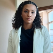 Natália posa, de pé, para a foto. É uma mulher jovem e negra, de cabelos até o ombro, trajando um blazer branco e cropped preto.