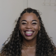 Agnes é uma mulher jovem e negra. Maquiada, ela sorri para a foto. Seus cabelos vão até abaixo do ombro