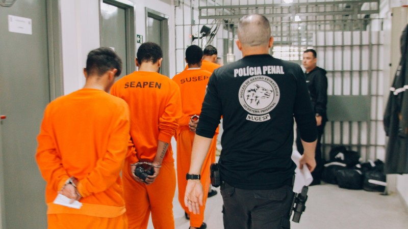 Presos, vestindo laranja, são conduzidos por um policial de roupa preta onde se lê Polícia Penal. Todos estão de costas, e os prisioneiros se dirigem para uma cela em fila indiana.