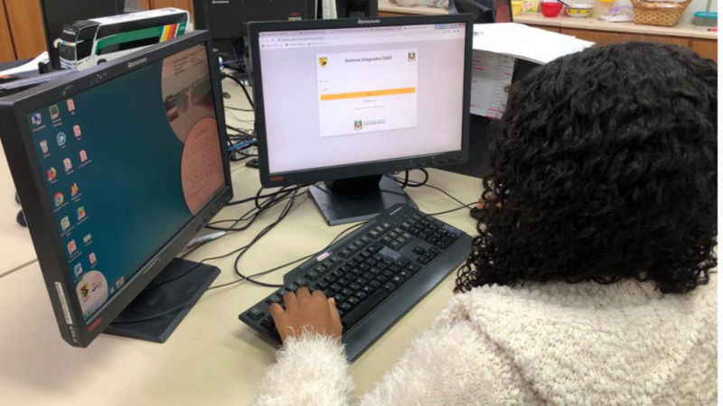Numa sala fechada, um mulher trabalha em frente a um computador com dois monitores