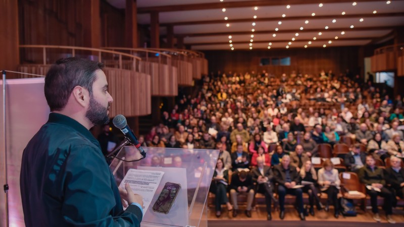 De lado, Gabriel aparece discursando para uma grande plateia, ao fundo, no teatro. Ele usa jaqueta verde-musgo