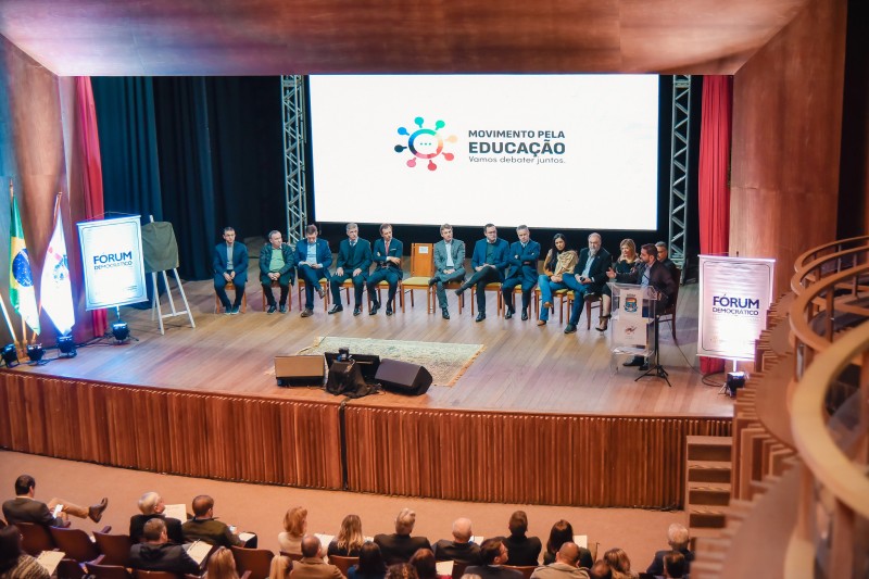 No palco de um anfiteatro, várias pessoas estão sentadas em cadeiras enfileiradas de frente para a plateia. Ao fundo, está um painel branco com a logo do Movimento pela educação