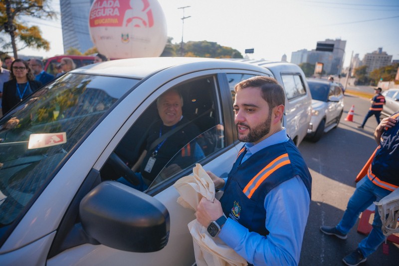 Gabriel Souza, trajando colete azul, sorri enquanto conversa com um motorista de carro pela janela do veículo