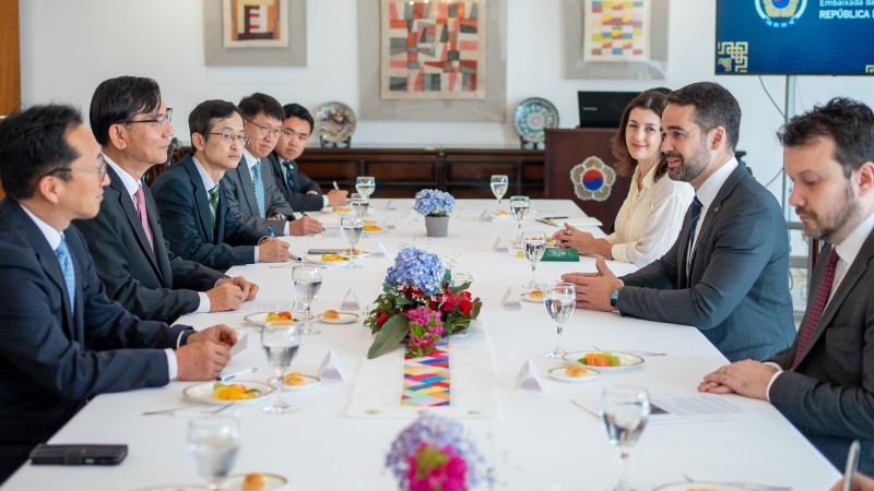 Os participantes estão sentados ao redor de uma mesa com toalha branca, pratos, copos e flores. À direita, estão Simone, Leite e Artur. Leite está sorrindo enquanto fala.