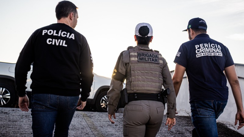 Dois policiais homens e uma policial mulher aparecem de costas. Nas camisas, est&aacute; escrito: Pol&iacute;cia Civil (policial da esquerda), Brigada Militar (do meio) e Per&iacute;cia Criminal (da direita)