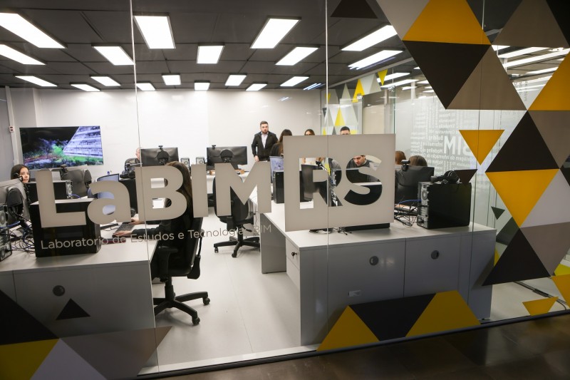 Imagem da divisória de vidro na qual está escrito LaBIM RS, atrás da qual podem ser vistas estações de trabalho com os computadores do laboratório e algumas pessoas conversando.